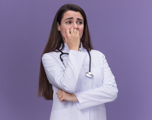 女焦急的年轻女医生穿着医用长袍 用听诊器咬指甲 一边看着紫色医疗听诊器焦虑