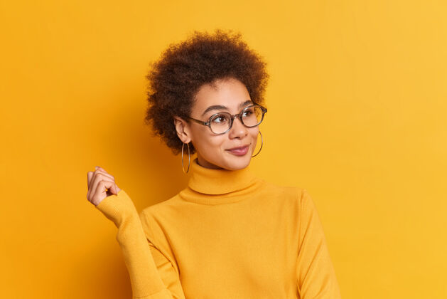高兴可爱的卷曲的非裔美国女学生戴着透明眼镜耳环和高领毛衣看着梦幻般的远方举起手来情感快乐黄色