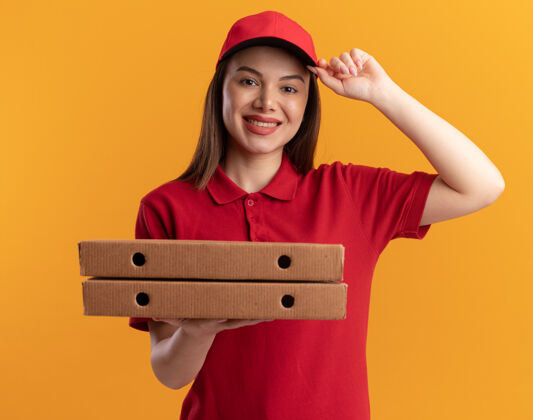漂亮身着制服的微笑漂亮的送货员把手放在帽子上 手里拿着橘黄色的比萨饼盒披萨手帽子