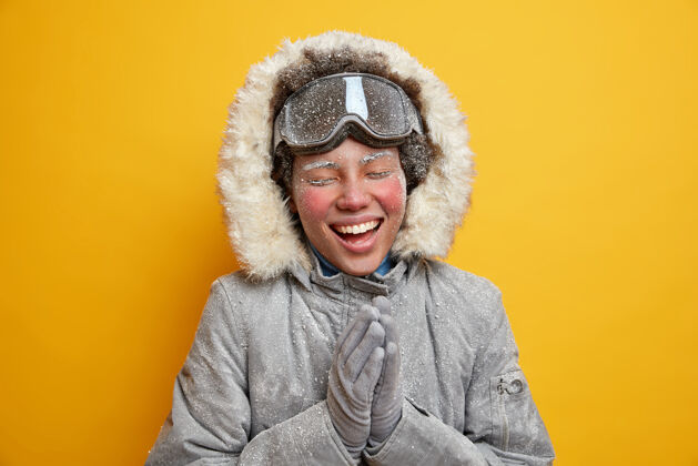 时尚美丽快乐的女子远足者喜欢冬天下雪 笑容灿烂 双手紧握 脸上覆盖着冰 闲暇时在山上穿着外套 戴着滑雪镜冬季假期活动夹克