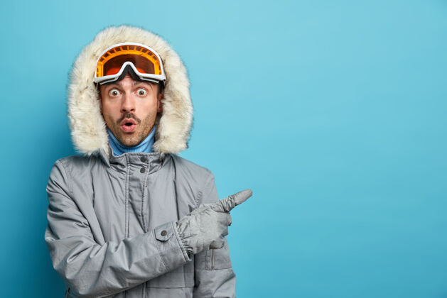 惊呆了活跃的惊呆了的人在冬天的山上训练去滑雪板穿灰色夹克和滑雪镜在空旷的空间用惊讶的表情表示夹克指示器惊艳