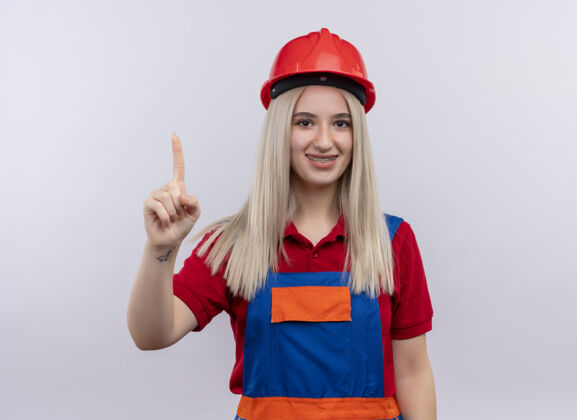 举起来微笑着的年轻金发工程师建筑工女孩穿着制服 戴着牙套 在孤立的空白处竖起手指年轻人牙套手指