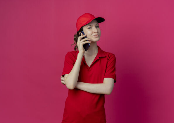 制服穿着红色制服 戴着帽子的年轻漂亮送货女孩站在那里 以封闭的姿势打电话 看着被隔离在深红色背景上的摄像头 还有复印空间封闭红色站立