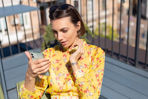 积极阳光明媚的夏日咖啡馆露台上穿着黄色衣服的女人拿着手机自拍打视频电话使用科技女性