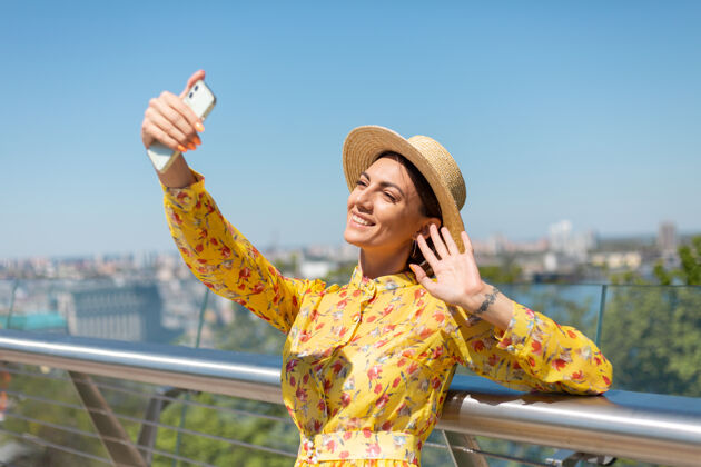 短信穿着黄色夏装戴着帽子的女人的户外肖像在电话里自拍 站在桥上 城市景色令人惊叹聊天表情模型
