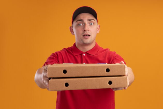 拿着身着制服 头戴鸭舌帽的年轻送货员高兴地把披萨盒放在隔离在橙色墙上的前面披萨脸请