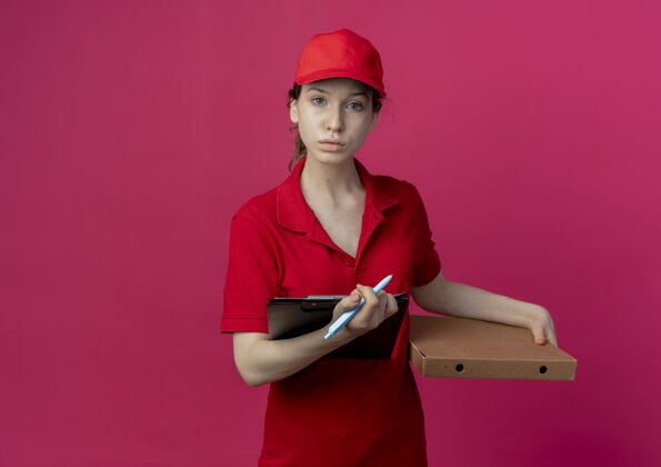 持有年轻漂亮的送货女孩 穿着红色制服 戴着帽子 手里拿着比萨饼包装笔和剪贴板 看着被隔离在深红色背景上的相机 还有复印空间深红色包装年轻