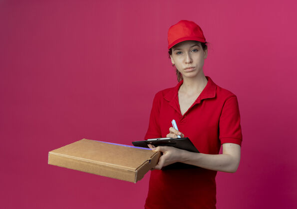 帽子自信的年轻漂亮的送货女孩 穿着红色制服 戴着帽子 手里拿着披萨包装笔和剪贴板 背景是深红色的披萨漂亮包装