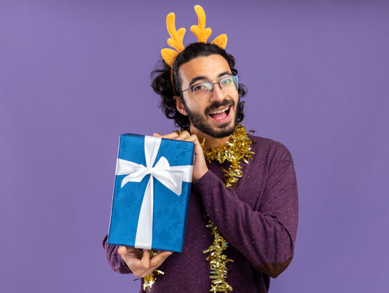 年轻快乐的年轻帅哥戴着圣诞发箍 脖子上戴着花环 手里拿着蓝色背景的礼品盒圣诞节花环盒子