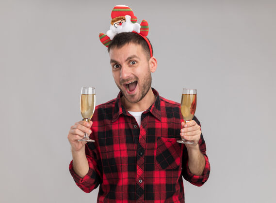 新年令人印象深刻的年轻白种人戴着圣诞头带拿着两杯香槟看着隔离在白色背景上的相机印象两个持有