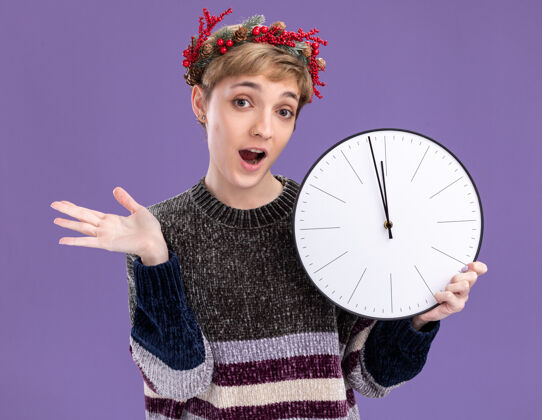花圈令人印象深刻的年轻漂亮女孩戴着圣诞花环手持时钟看着相机显示空手孤立在紫色的背景新年手圣诞