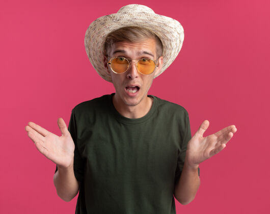 人愤怒的年轻帅哥 穿着绿色衬衫 戴着眼镜 戴着帽子 两手分开放在粉红色的墙上穿着帽子手