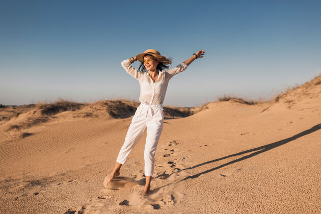 冒险时髦的微笑美丽的快乐女人穿着白色的衣服在沙漠中奔跑和跳跃在日落时戴着草帽旅游女人旅游