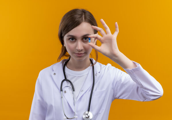 自信自信的年轻女医生穿着医用长袍和听诊器在孤立的橙色空间展示医用胶囊女性橙色医疗