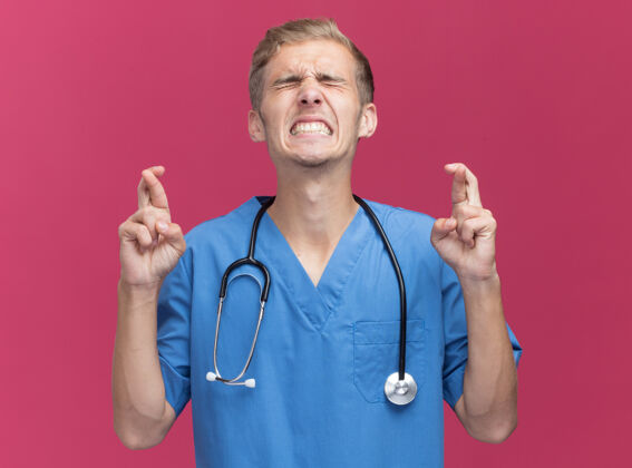 佩戴关注闭着眼睛的年轻男医生穿着医生制服用听诊器交叉手指隔离在粉红色的墙上医生站立脸