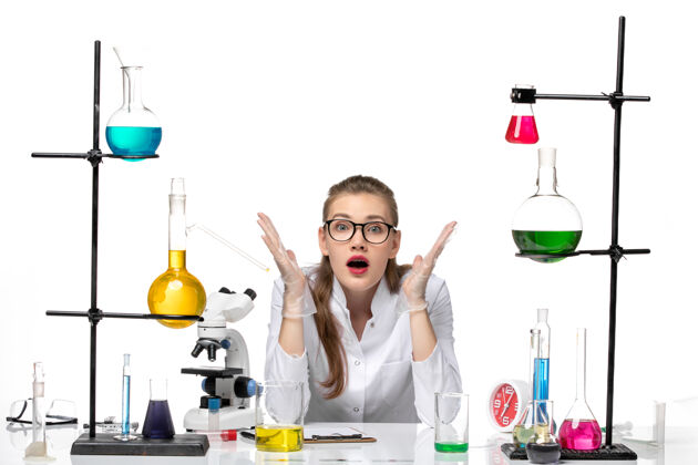 白色前视图身着医疗服的女化学家在桌子前拿着白色背景上的溶液化学大流行的冠状病毒视图玻璃桌子