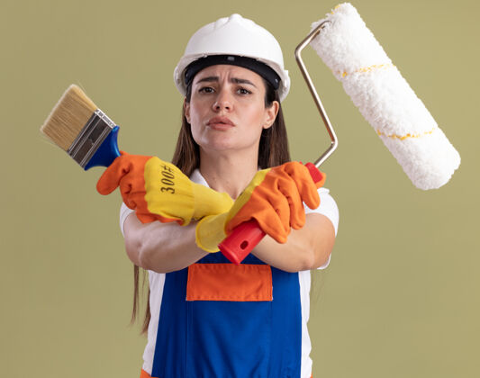 橄榄穿着制服 戴着手套的严格的年轻的建筑工人妇女拿着油漆刷和过路刷隔离在橄榄绿的墙上建设者交叉严格