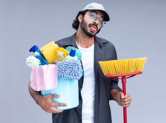 年轻快乐的年轻帅气的清洁工 穿着t恤 戴着帽子 拿着一桶清洁工具 露出舌头 拖把被隔离在白色的墙上帅哥家伙帽子