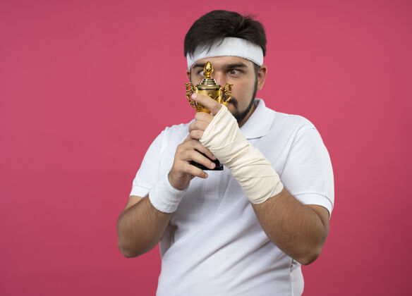 运动年轻的运动型男子戴着头带和腕带 手腕缠着绷带 拿着冠军杯 看着隔离在粉红色墙上的复制空间持有头带冠军