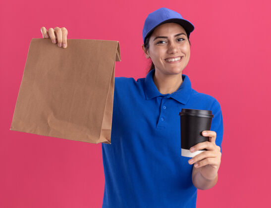 咖啡带着微笑的年轻送货女孩 穿着制服 戴着帽子 手里拿着一个纸食品包 粉红色的墙上隔着一杯咖啡包装年轻人送货