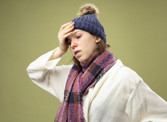 疾病咳嗽的年轻生病的女孩一边看一边穿着白色长袍 戴着冬天的帽子 戴着围巾 手放在额头上 隔离在橄榄绿上额头年轻围巾