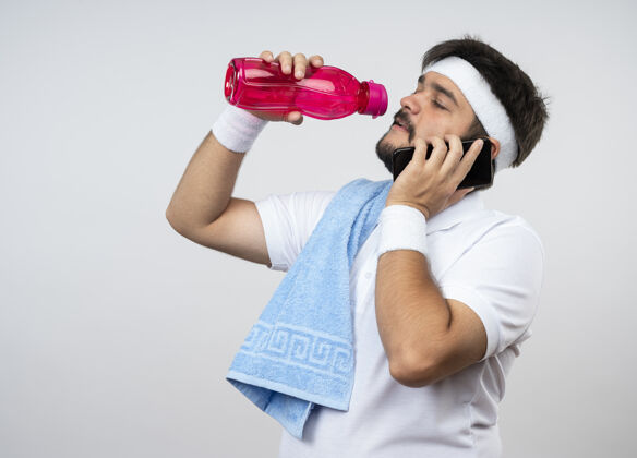 运动疲惫的年轻人戴着头带和手镯 肩上带着毛巾 从隔离在白色墙壁上的水瓶里喝水 并留有复制空间穿肩膀头带
