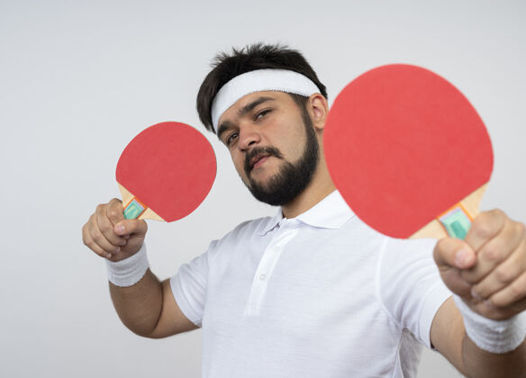 运动自信的年轻人戴着头带和腕带 举着乒乓球拍孤立地站在白墙上腕带头带抱着