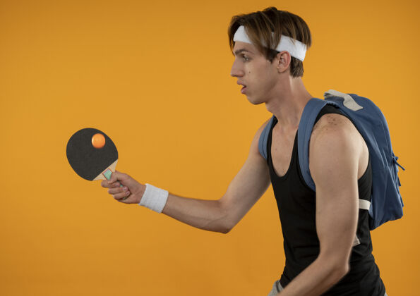 年轻站在侧视图年轻的运动家伙穿着背包与头带和手环举行乒乓球拍隔离在橙色墙上穿立场运动