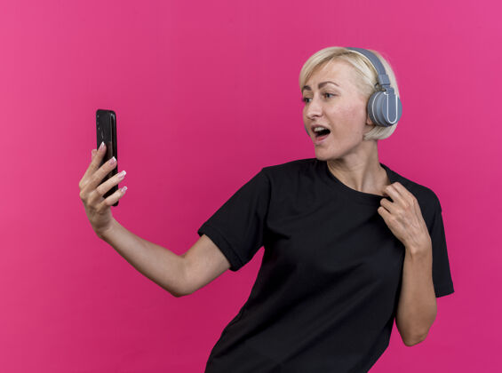 抓取令人印象深刻的中年金发斯拉夫妇女戴着耳机拿着手机看着她的t恤衫孤立在粉红色的墙上人脸耳机