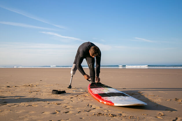 皮带冲浪者穿着潜水服和假肢 把冲浪板绑在沙滩上的脚踝上爱好板冲浪者