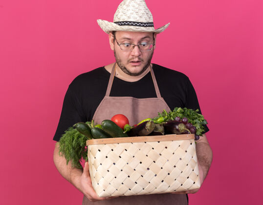 花园年轻的男园丁惊讶地戴着园艺帽 手里拿着一个孤立在粉红色墙上的菜篮子蔬菜园艺帽子