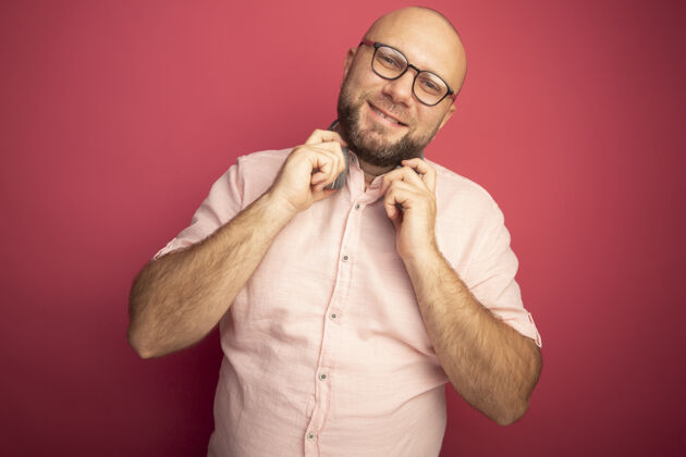 秃头微笑着歪着头的中年秃头男人穿着粉色t恤 脖子上戴着眼镜和耳机 隔离在粉色的墙上男人微笑倾斜