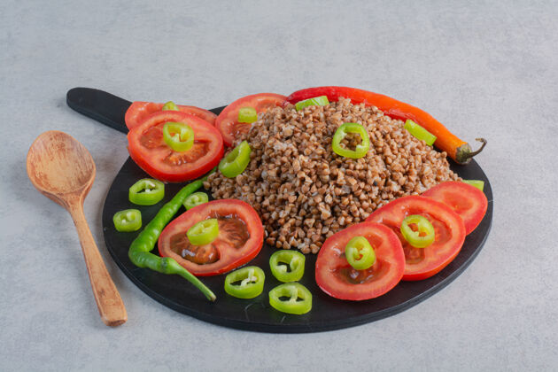 谷物在大理石表面的勺子旁边的木板上放着一堆荞麦 上面撒着胡椒和西红柿胡椒配料天然