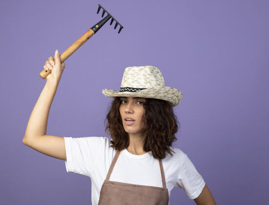 年轻自信的年轻女园丁穿着制服戴着园艺帽举起耙子人养紫色