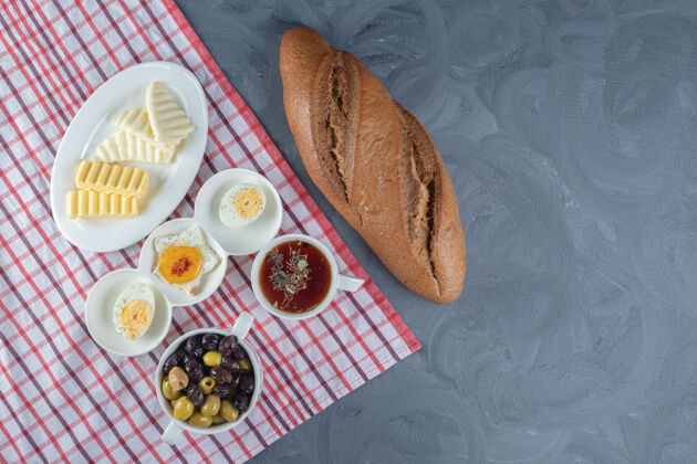 奶酪桌布下面是早餐包面包和一盘盘奶酪 黄油和鸡蛋 大理石背景上是茶和橄榄橄榄杯子面包