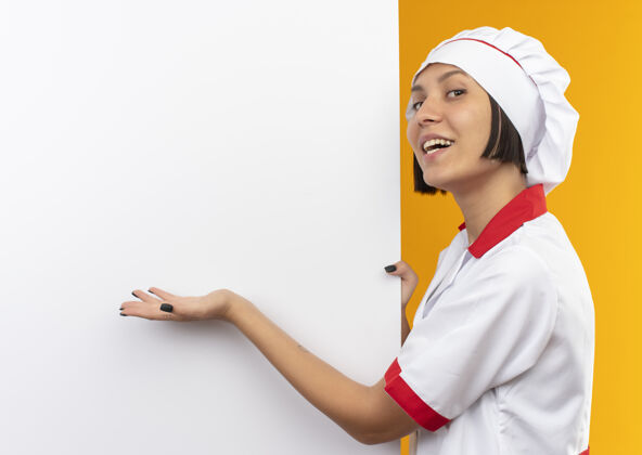 指着身着厨师制服的年轻快乐的女厨师站在白色的墙壁旁 手拿着并指着隔离在橘黄色上的墙烹饪靠近站着