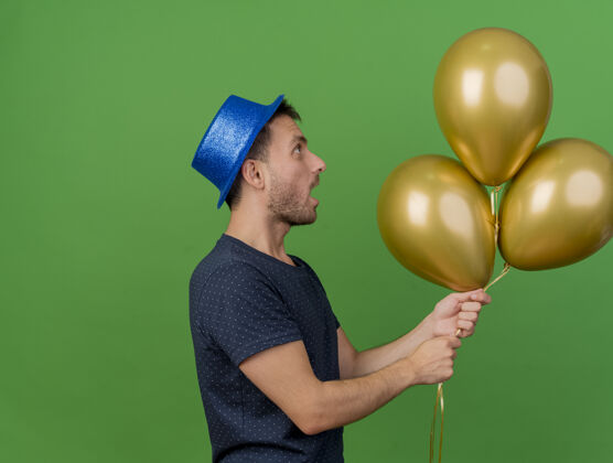 气球一个戴着蓝色派对帽的帅哥站在一边看着 手里拿着氦气球 这些气球被隔离在绿色的墙上 留着复制空间人帽子聚会