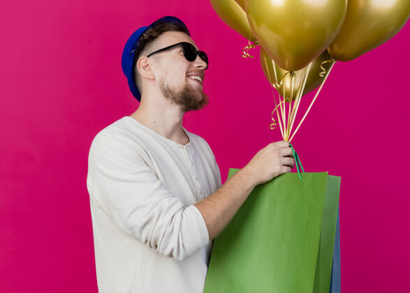 立场面带微笑的年轻帅哥斯拉夫党人戴着党帽和太阳镜站在侧视图看直手持气球和纸袋孤立在粉红色的墙上斯拉夫黄金帽子