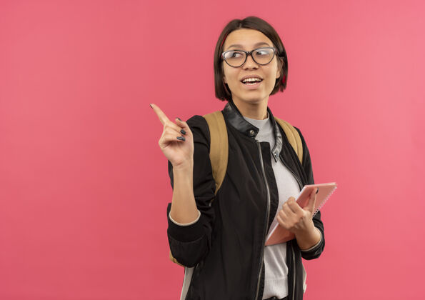 笔记快乐的年轻女学生戴着眼镜 背着书包 拿着便签本 指着粉色的一边看着孤立的指壁板学生