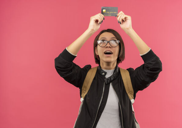 信用印象深刻的年轻女学生戴着眼镜 背着书包 看着信用卡上的粉红色隔离佩戴年轻眼镜