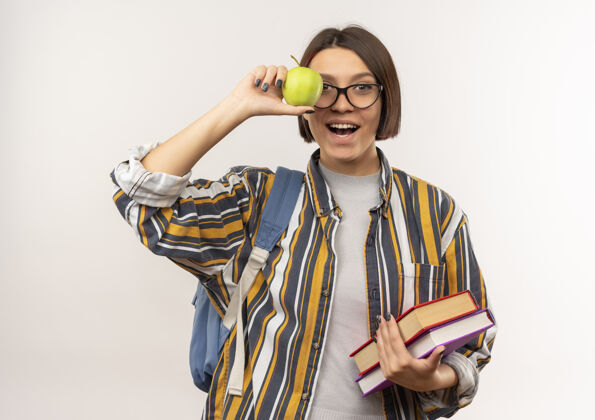 年轻快乐的年轻女学生戴着眼镜 背着书包 手里拿着苹果 孤零零地站在白色的地板上戴学生背