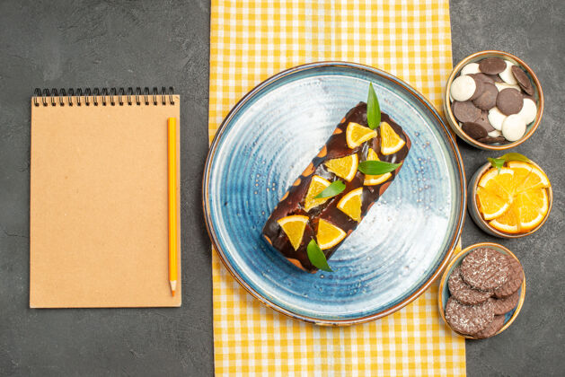 杯子黄色毛巾上的美味蛋糕和黑桌子上笔记本旁的饼干蔬菜碗玻璃