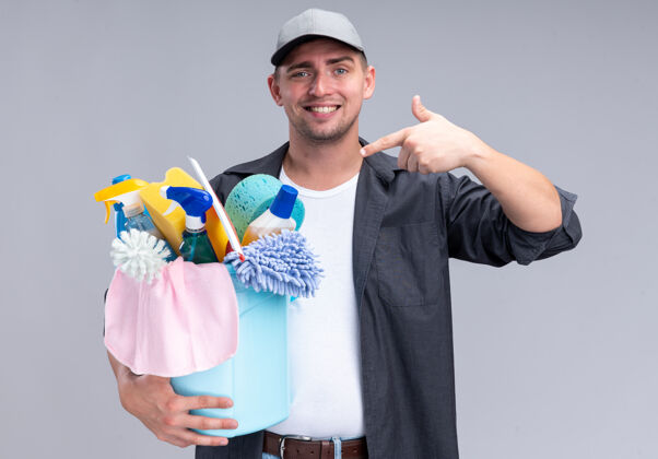 家伙微笑着的年轻帅气的清洁工穿着t恤 戴着帽子 指着隔离在白墙上的一桶清洁工具T恤微笑帽子