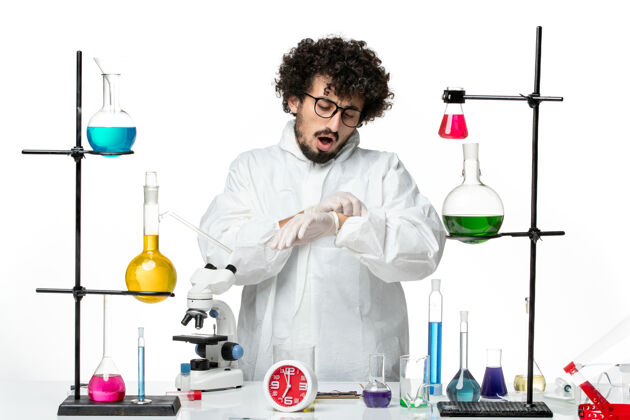 套装前视图身穿白色特殊套装的年轻男性科学家站在桌子旁 用解决方案看着他的手腕烧杯溶液视图