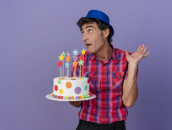 蛋糕令人印象深刻的中年白种人聚会男子戴着聚会帽拿着生日蛋糕抬头看显示空手孤立在紫色背景与复制空间手中年复制