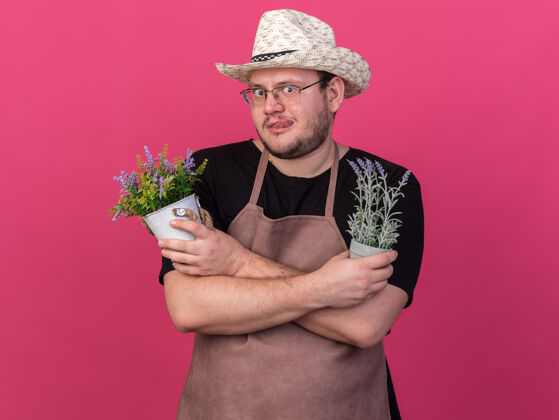 穿可疑的年轻男园丁戴着园艺帽 拿着和粉红色墙上隔离的花盆里的花交叉交叉帽子可疑