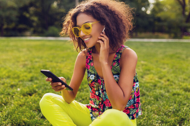 音乐年轻时尚微笑的黑人女子用智能手机听音乐无线耳机在公园里玩得开心 夏日时尚多彩的风格 坐在草地上 戴着黄色太阳镜兴奋太阳镜年轻