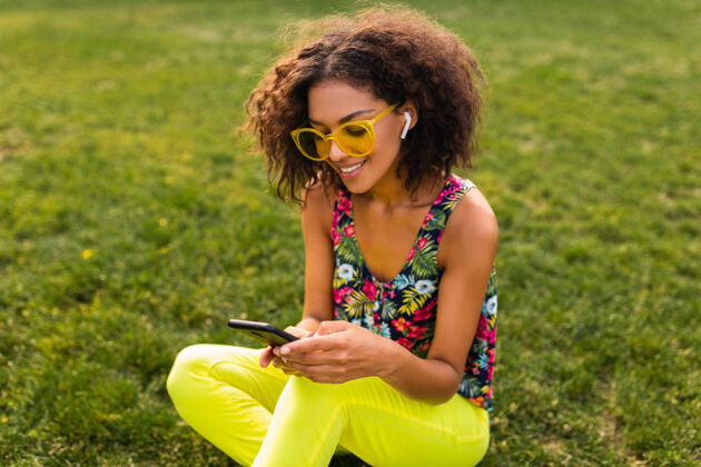 女士年轻时尚微笑的黑人女子使用智能手机 通过无线耳机听音乐 在公园里玩得很开心眼镜欢呼太阳镜