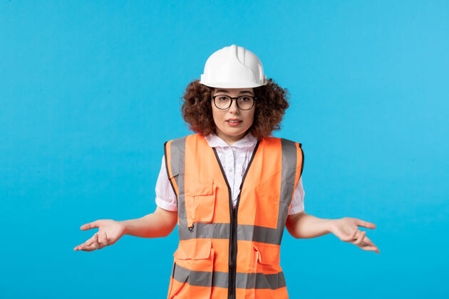 专业蓝色墙壁上穿着制服戴着头盔的困惑的女建筑工人的正视图成人制服承包商
