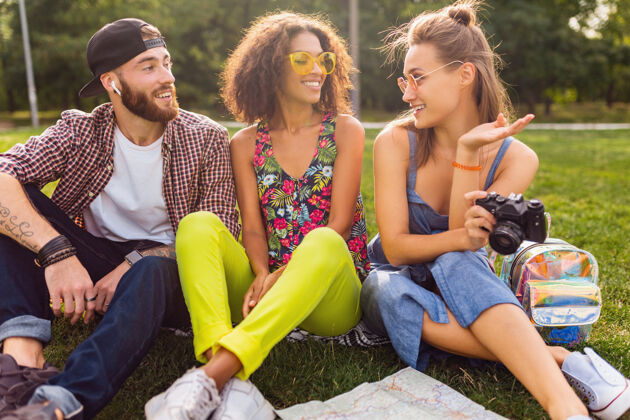 女人快乐的年轻朋友陪伴坐在公园里 男女同乐 缤纷的夏日潮人时尚风格 带着相机旅行 谈笑风生太阳镜相机团体